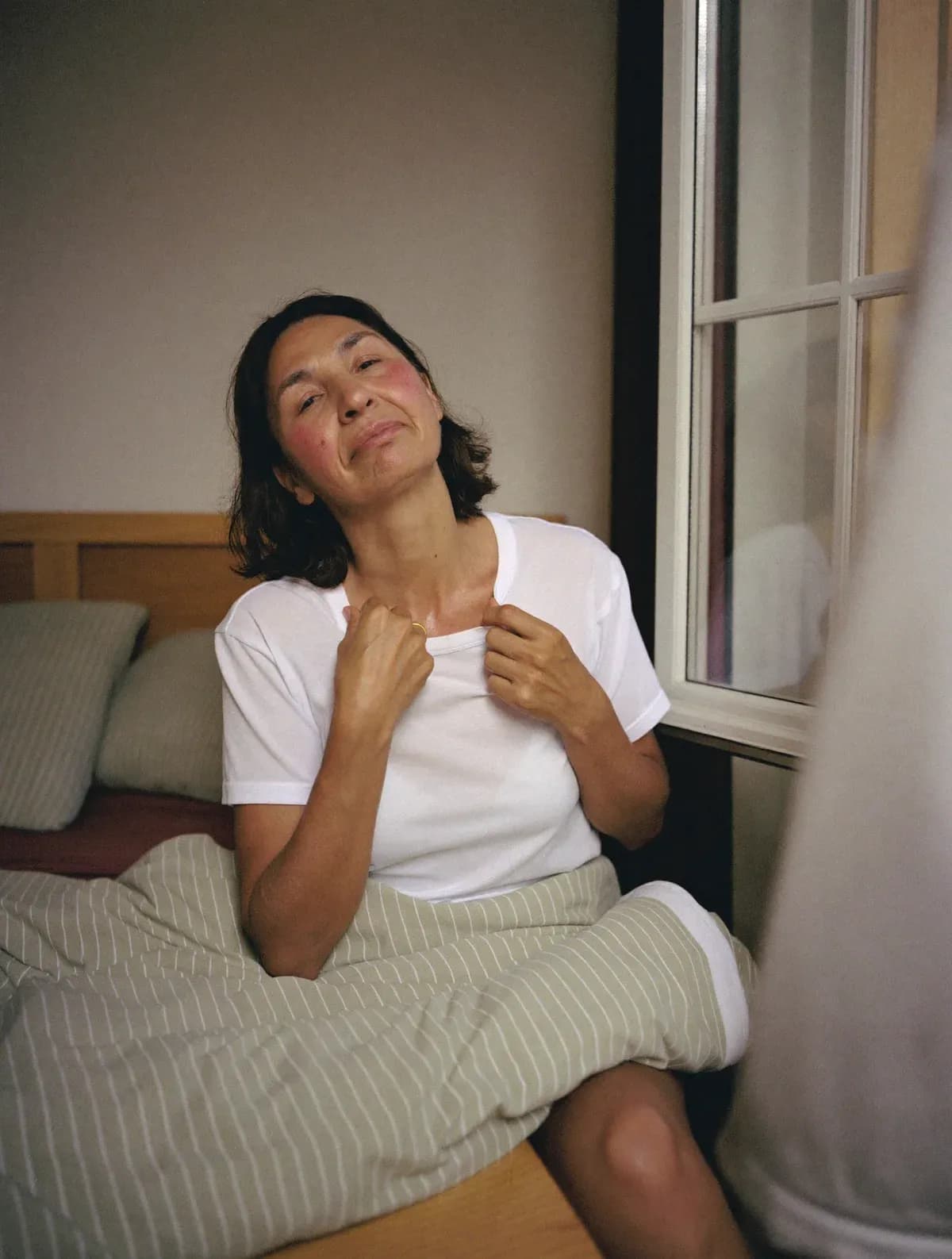 Woman sitting up in bed by an open window, cheeks flushed, pulling her t-shirt collar away from her neck
