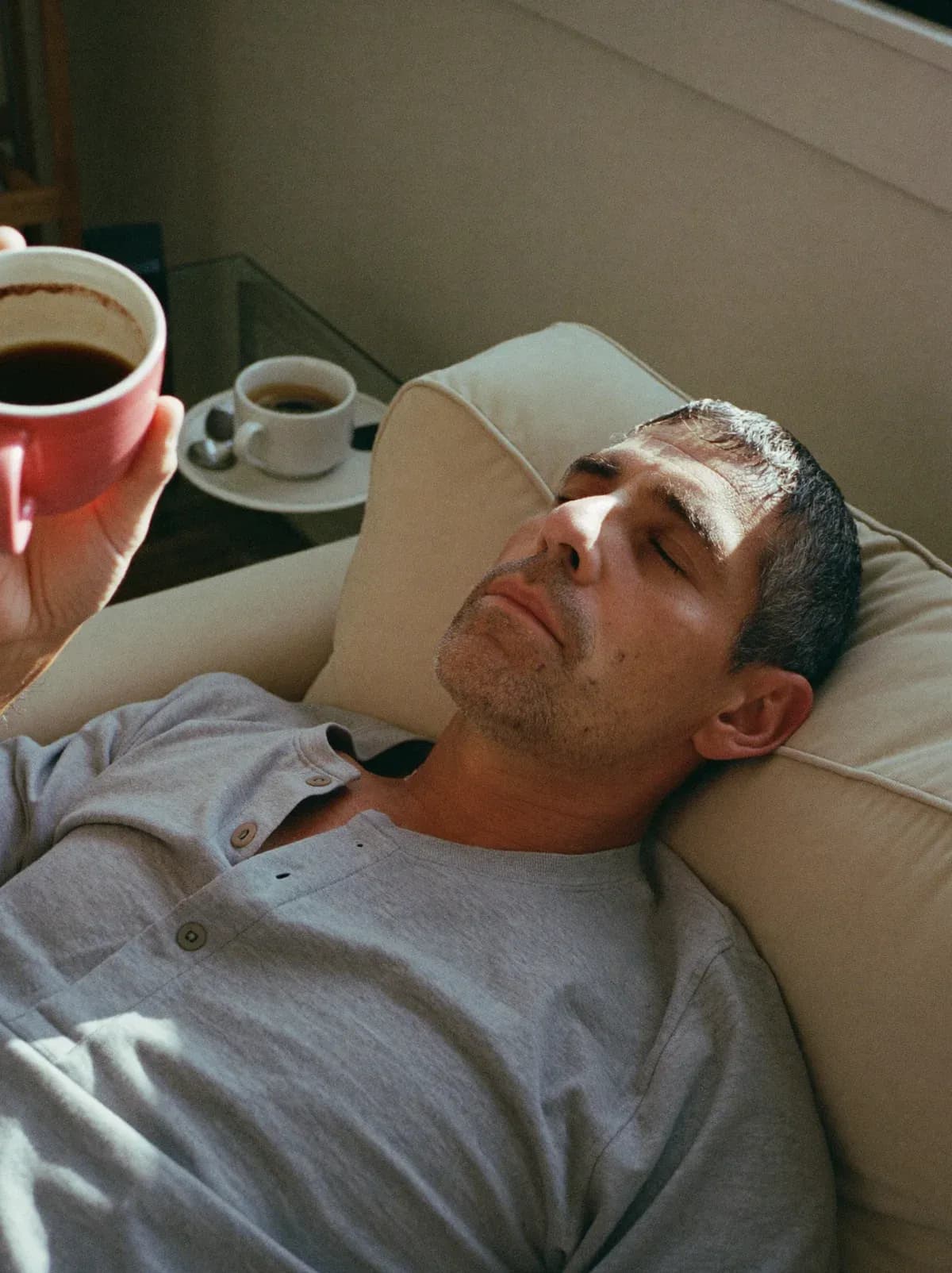 Tired man resting his head back on the sofa, eyes closed, a coffee in his hand